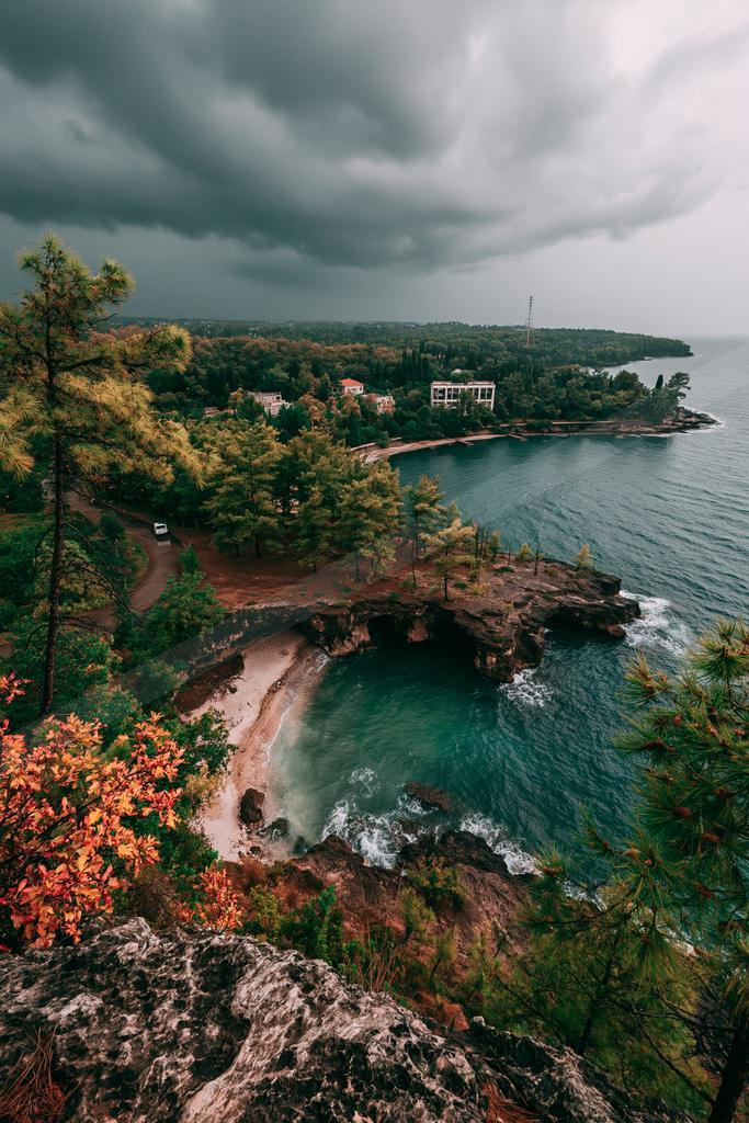 High-angle view of a rocky coastline with turquoise water, a small sandy beach in a cove, dense green and autumn-colored forest, buildings along the shore, and