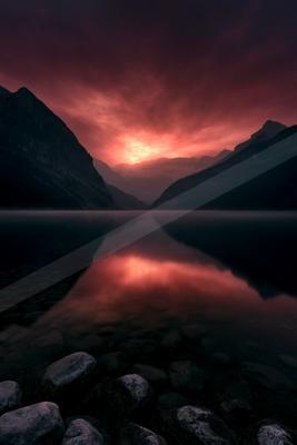 A dramatic red and purple sunset illuminates the sky above a calm mountain lake, perfectly reflected in the still water with a rocky foreground shore.