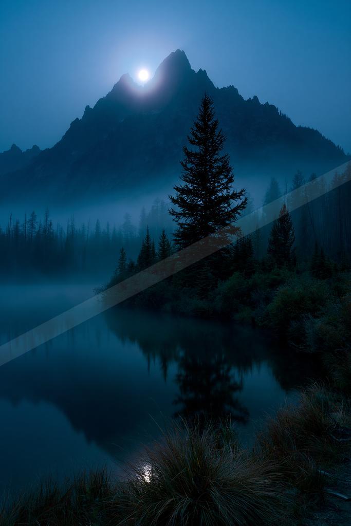 A bright moon illuminates a jagged mountain peak above a misty lake surrounded by dark pine trees, with the scene perfectly reflected in the calm water at night