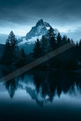 A serene lake mirrors a snow-capped mountain and surrounding evergreen trees under a twilight sky.