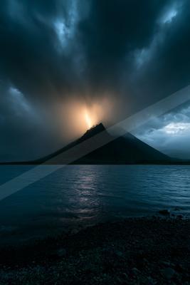 A dark conical volcano rises behind a rippling lake under a stormy sky, with a bright lightning bolt striking the summit amid heavy clouds.