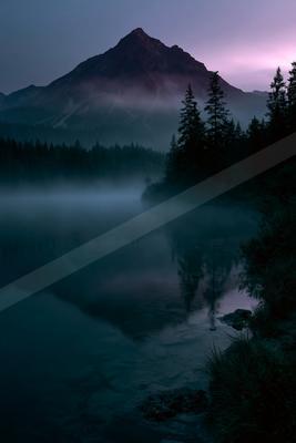 A serene mountain lake shrouded in thick fog at twilight, with the peak of a rugged mountain rising above the mist and the calm water reflecting the dark silhou