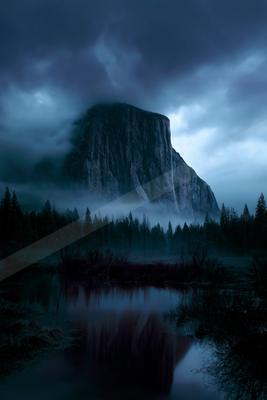 A massive rocky mountain peak rises through thick blue-grey fog above a dense coniferous forest, its reflection visible in a calm foreground lake under a stormy