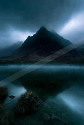 A dramatic, mist-covered mountain peak is perfectly reflected in the still surface of a lake, with foreground rocks and grass under a moody, overcast sky.