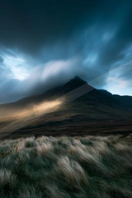 A rugged mountain peak shrouded in atmospheric clouds, illuminated by soft light against a dark sky.