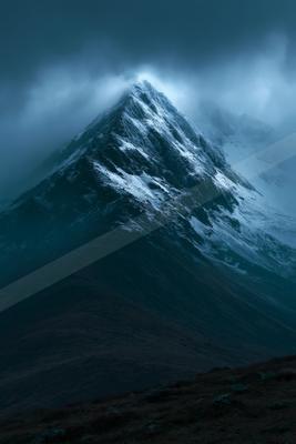 A sharp, snow-covered mountain peak rises through thick, swirling clouds under a dark, moody sky, with a foreground of dark, barren hills.