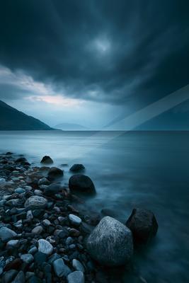 A pebble-strewn lakeshore with large rocks in the foreground meets a calm, smooth lake under a dark, dramatic stormy sky with distant mountains visible on the h