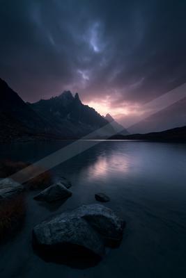 A serene alpine lake reflects the dramatic twilight sky and jagged mountain peaks under dark, moody clouds, with large rocks and grasses in the foreground.