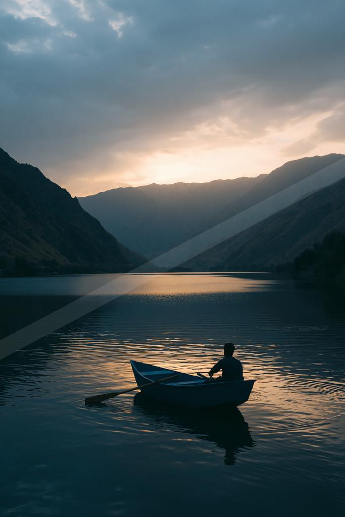 A silhouetted man rows a small boat across a calm mountain lake as the sun sets behind distant peaks, casting golden light and reflections on the water.