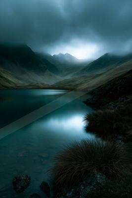 A serene mountain lake with calm reflective water surrounded by rugged hills and shrouded in thick mist under a dramatic cloudy sky.