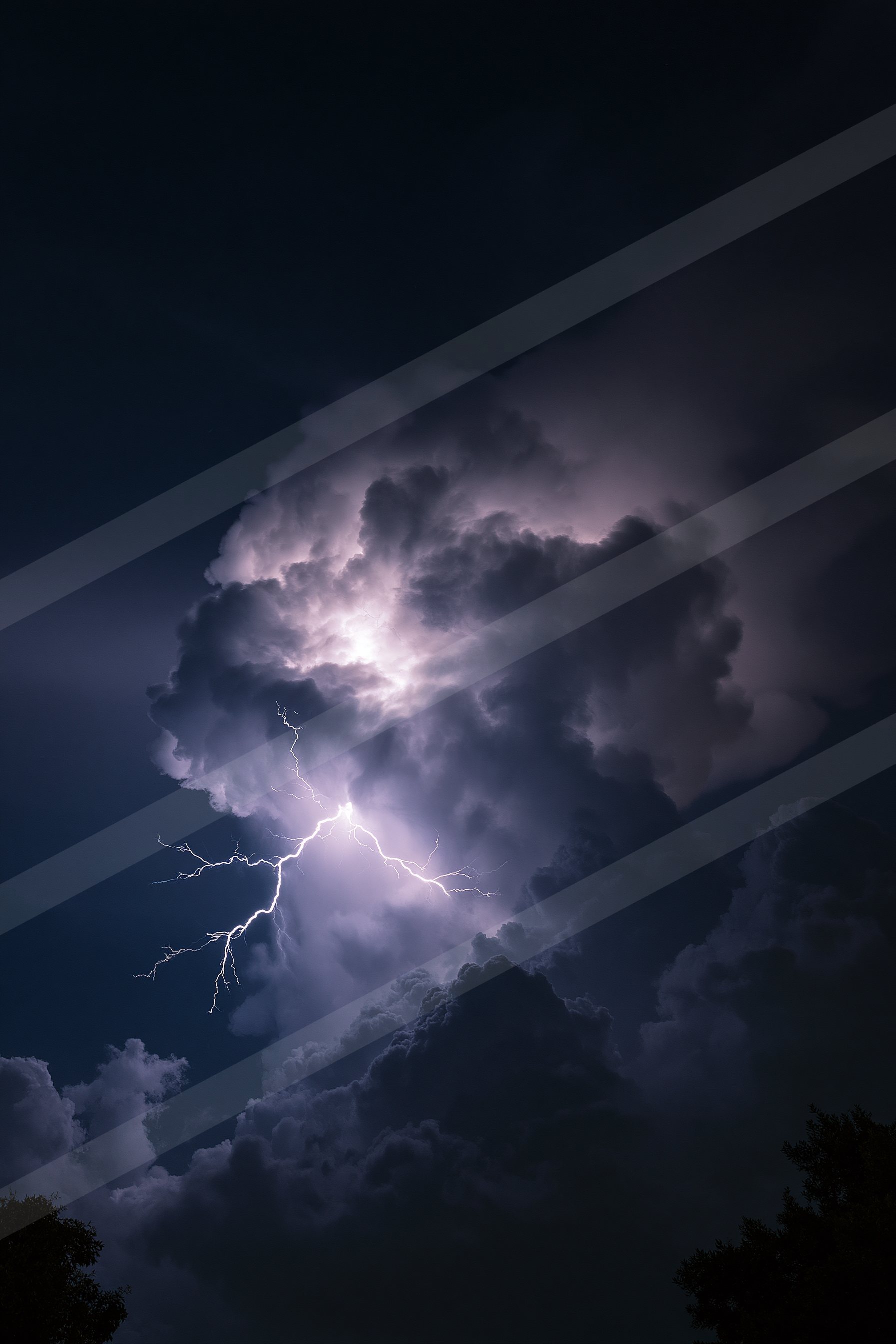 A powerful lightning bolt illuminates dark storm clouds against a deep blue night sky, with silhouettes of trees visible below.