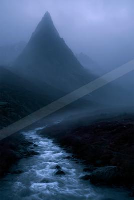 A fast-flowing stream winds through a rocky, vegetated valley toward a tall, sharply peaked mountain shrouded in thick blue-gray fog.