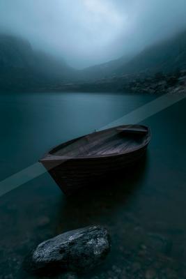 A dark wooden rowboat floats on the calm water of a misty mountain lake, with rocky shores and fog-covered peaks in the background.