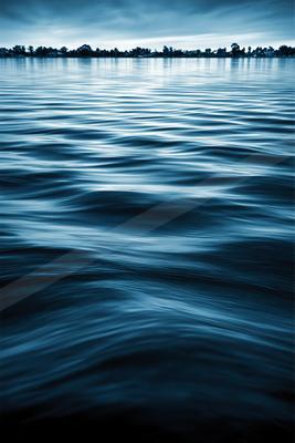 Calm blue water with gentle ripples reflecting the overcast sky, distant tree-lined shore visible on the horizon