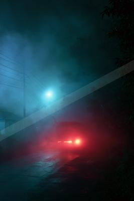 A car with glowing red taillights drives away down a wet rural road enveloped in thick blue and teal fog at night, with a bright streetlight and utility poles v