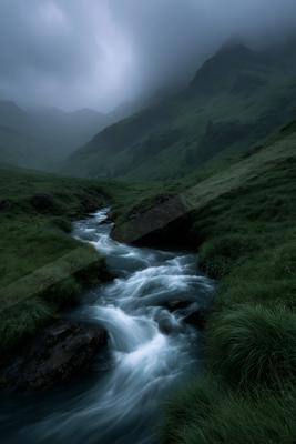 A fast-flowing stream with white water rushes through a lush green valley with rocks and grass, surrounded by misty mountains under a cloudy sky.
