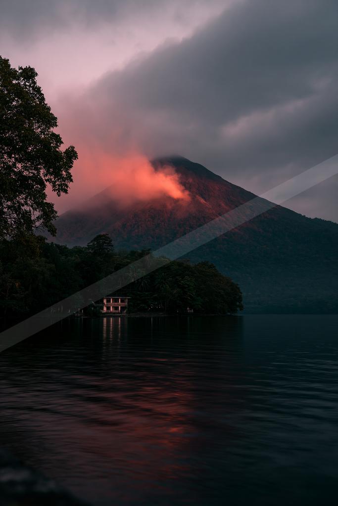 A dramatic volcano emits a red glow through low clouds at dusk, reflected in a calm lake with a forested shoreline and a small building in the foreground.