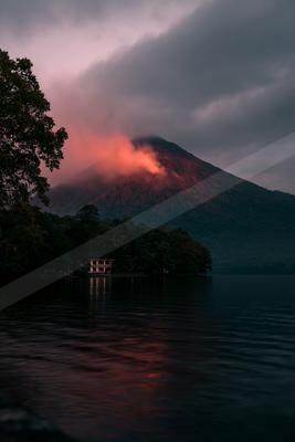 A dramatic volcano emits a red glow through low clouds at dusk, reflected in a calm lake with a forested shoreline and a small building in the foreground.