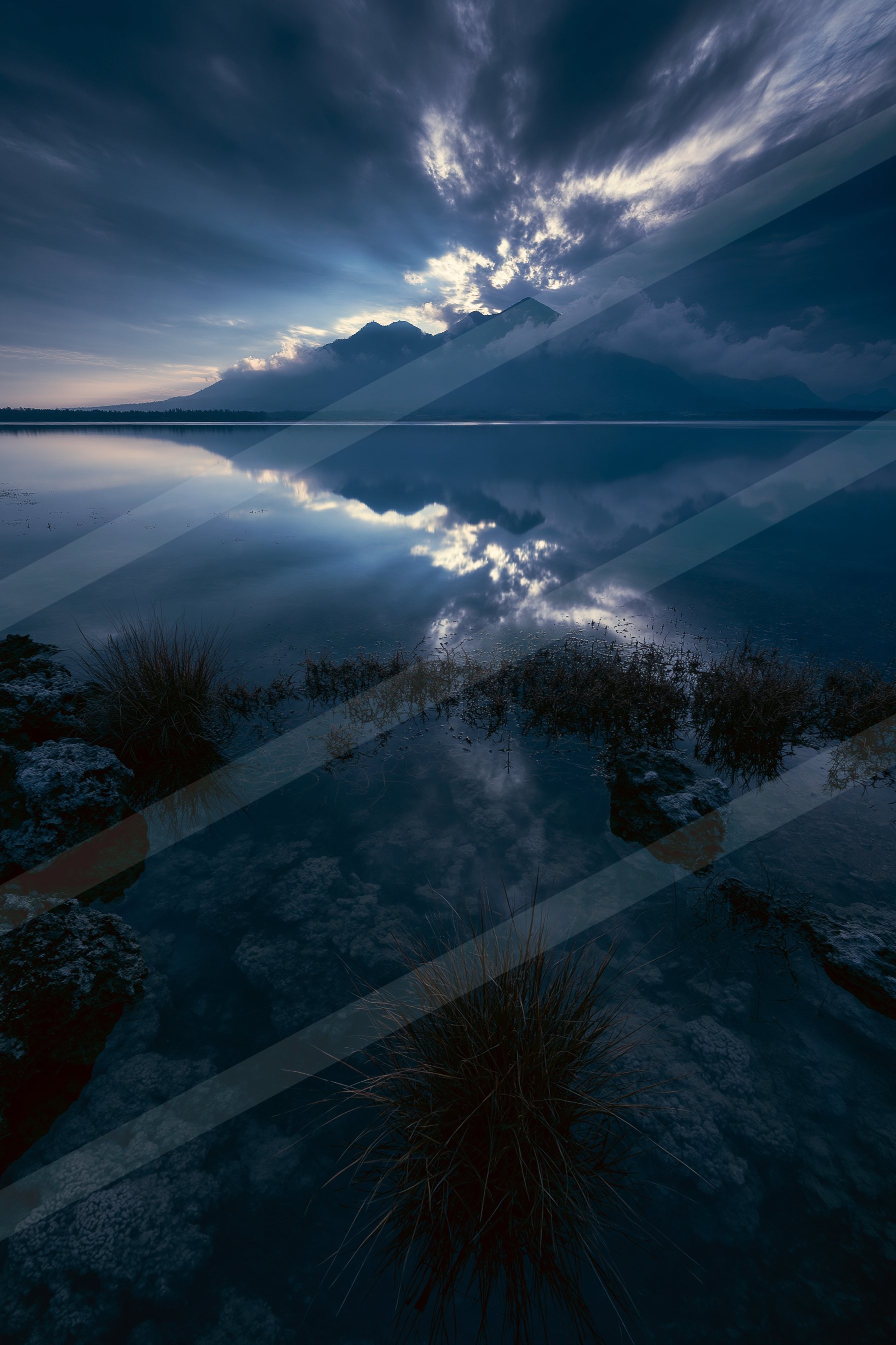 A serene lake reflects a dramatic mountain silhouette under a moody twilight sky filled with clouds.