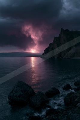 A dramatic pink and purple sunset glows through dark storm clouds, casting a vibrant reflection on a calm lake beside a steep rocky mountain and foreground boul