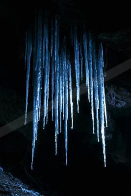 Long, glowing blue icicles hanging from the ceiling of a dark cave, illuminated dramatically against the black rock background.