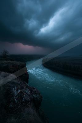 A powerful river flows through a rocky canyon under a sky filled with dark, ominous storm clouds, creating a moody and atmospheric landscape.