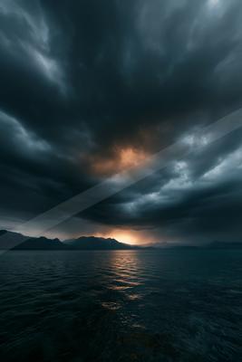 Moody dark storm clouds fill the sky above a mountain range and a large body of water, with a warm orange glow from the setting sun breaking through the clouds 