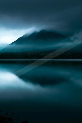 A serene lake reflects the silhouette of a mountain shrouded in mist against a dark, brooding sky.