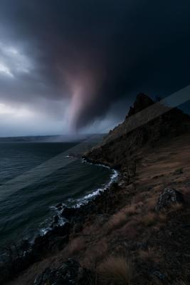 A powerful waterspout descends from dark storm clouds over a large body of water, next to a rocky, grassy shoreline with sparse trees under a moody sky.