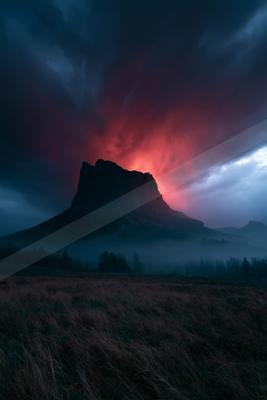A silhouetted rocky mountain peak rises above a layer of fog and a field of dry grass under a dramatic sky glowing with intense red and dark blue clouds.