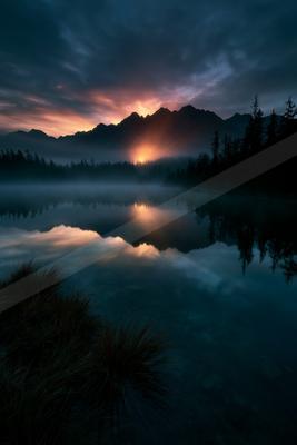 A dramatic sunrise with orange and pink light breaking through clouds behind silhouetted mountains, perfectly reflected in a calm misty lake surrounded by dark 