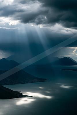 Crepuscular rays pierce through dark storm clouds, illuminating a vast mountain lake surrounded by rugged peaks.