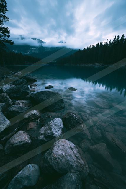 A serene lake surrounded by a rocky shoreline, reflecting the surrounding trees and distant mountains under a cloudy sky.