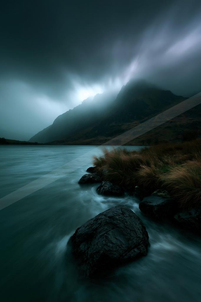 A dark, atmospheric landscape featuring a calm lake with smooth water from long exposure, rocky shoreline with dry grass, and a large mountain partially obscure