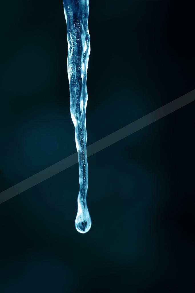 A translucent blue icicle hangs vertically against a dark blurred background with a single water droplet forming at its tip.