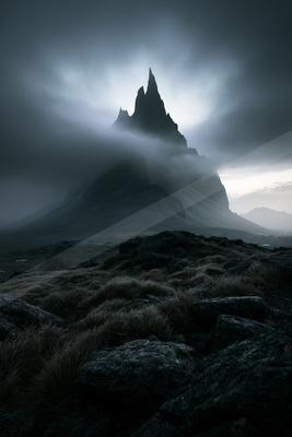 A dramatic dark mountain peak emerges from swirling mist against a moody sky, creating an otherworldly atmosphere.