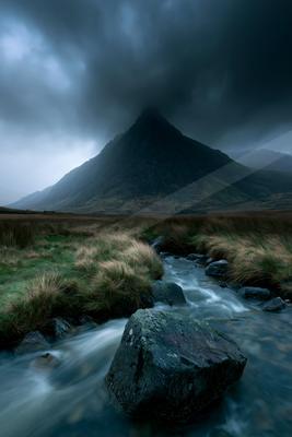 A fast-flowing stream with large rocks winds through a grassy moorland toward a dark, mist-covered mountain peak beneath heavy storm clouds.