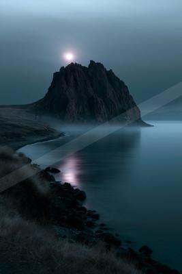 A large dark rocky island rises from a calm sea under a bright full moon, with thick fog at its base and a rocky grassy shoreline in the foreground.