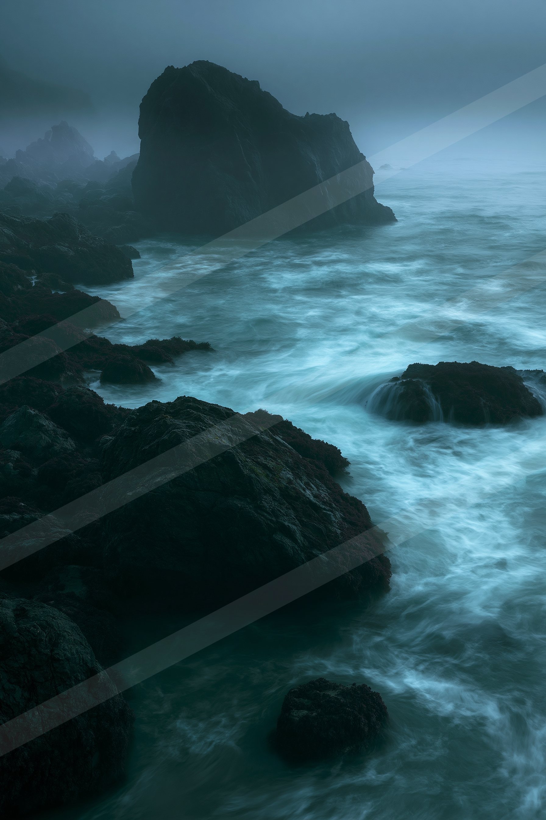 A moody coastal landscape featuring jagged rocks, turbulent ocean waves, and a misty atmosphere during dusk.