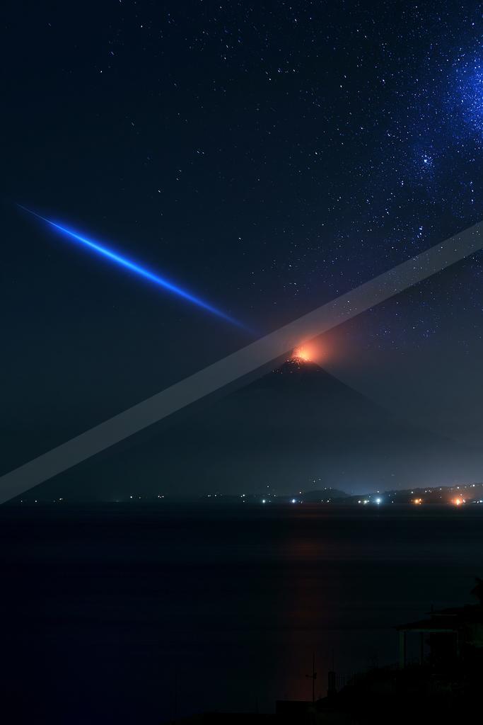 A volcano erupting with glowing lava under a star-filled night sky, featuring a prominent blue star trail and reflections on the water below.