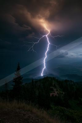 A powerful lightning bolt illuminates the night sky over a dark forested mountain range, creating a striking contrast against the stormy clouds.