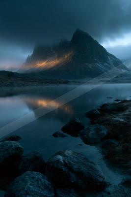 A sharp, rocky mountain peak illuminated by warm light breaks through heavy dark clouds, its reflection mirrored perfectly in the calm surface of a foreground l