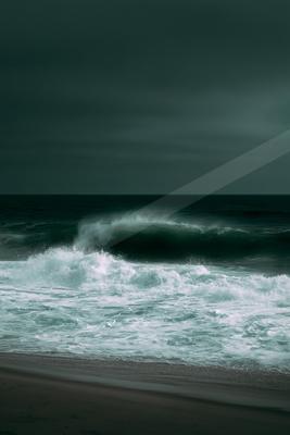 A powerful ocean wave crashes with white foam onto a dark sandy beach under a moody, overcast sky.