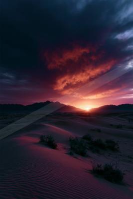 Vast sand dunes with sparse desert shrubs under a dramatic sky filled with fiery red, orange, and dark purple clouds at sunset, with the sun visible on the hori