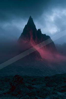 A sharp rocky mountain peak illuminated with red light rises through thick fog under a dark cloudy sky, with a rocky foreground.