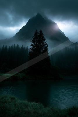 A tall dark coniferous tree stands in the foreground beside a calm lake, with a massive misty mountain peak rising behind a dense forest under brooding clouds.