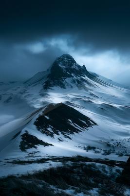 A rugged, snow-covered mountain peak rises sharply into a dark, cloudy sky with mist and dramatic lighting.