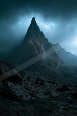 A sharp, rocky mountain peak rises dramatically through thick, dark clouds and mist, with a rugged, rocky foreground under a moody, overcast sky.