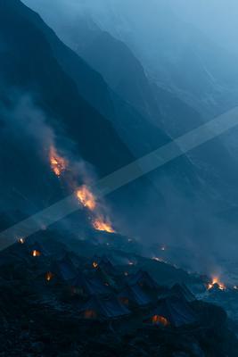 A campsite of blue tents illuminated by warm light, set against a backdrop of steep, snow-covered mountains. A fiery avalanche descends the slopes, casting an e