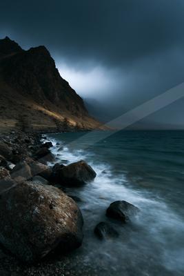 Moody landscape of a rocky lakeshore with large boulders in the foreground, a steep mountain slope on the left, and dark stormy clouds overhead with mist in the